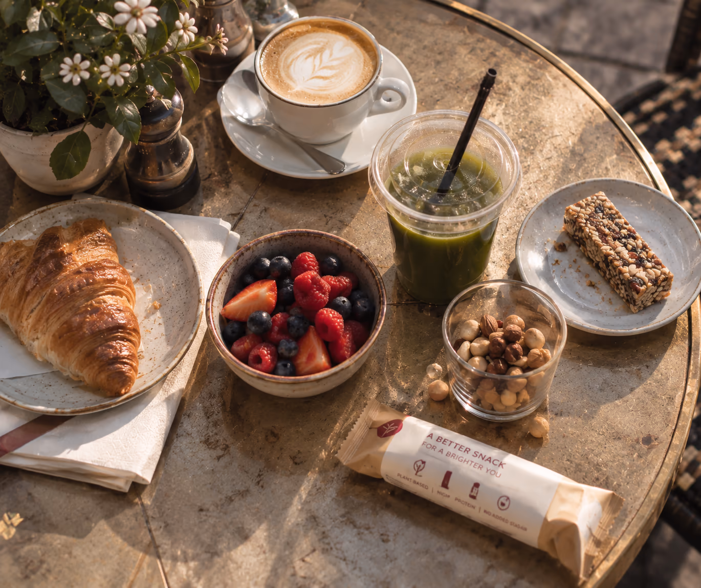 Frukost på ett kafébord med croissant, bär, grön smoothie, nötter, kaffe och en mellanmålsbar.